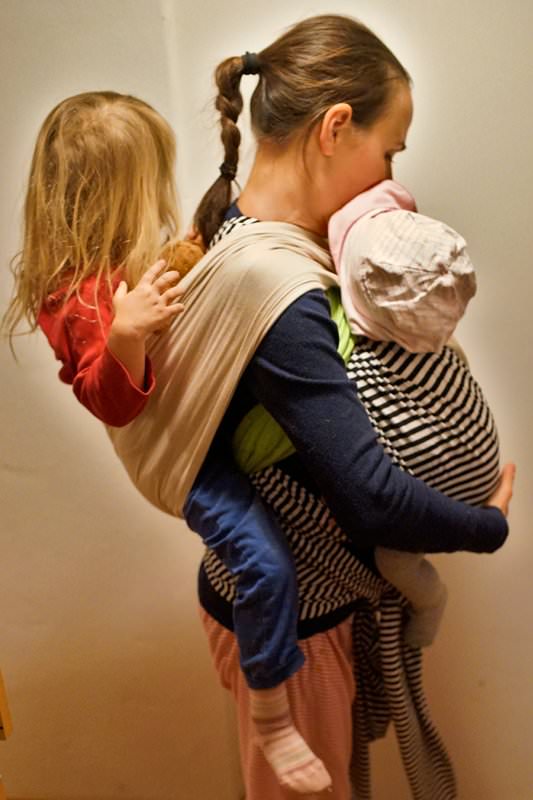ein Kind im Babytuch auf dem Rücken, ein Kind im elastischen Tragetuch auf dem Bauch