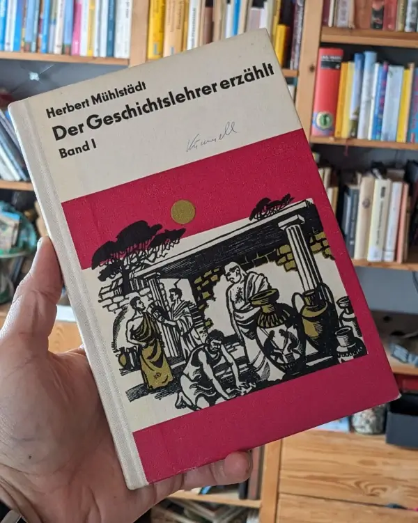 Buchcover von „Der Geschichtslehrer erzählt – Band I“ von Herbert Mühlstädt mit gezeichnetem antiken Szenenbild und handschriftlicher Signatur, im Hintergrund ein Bücherregal.