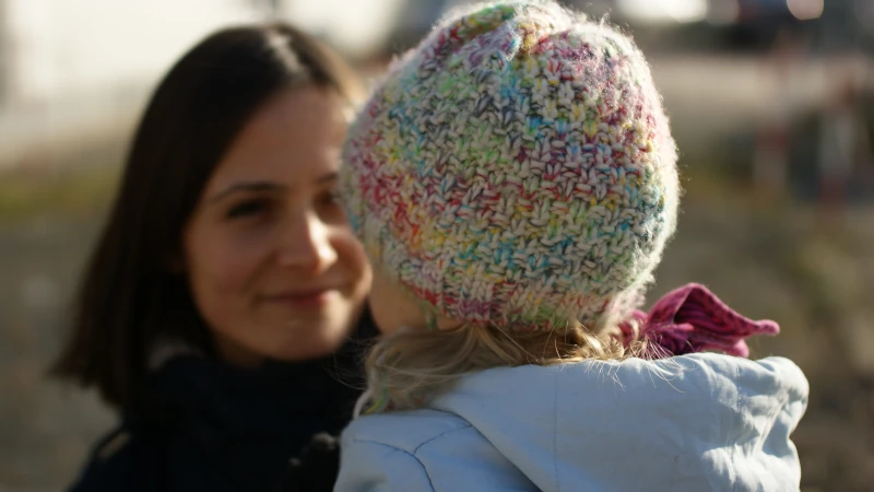 Eine Mutter (Evelin) schaut ihr Kind liebevoll an. Das Kind trägt eine bunte Wollmütze und ist von hinten zu sehen.