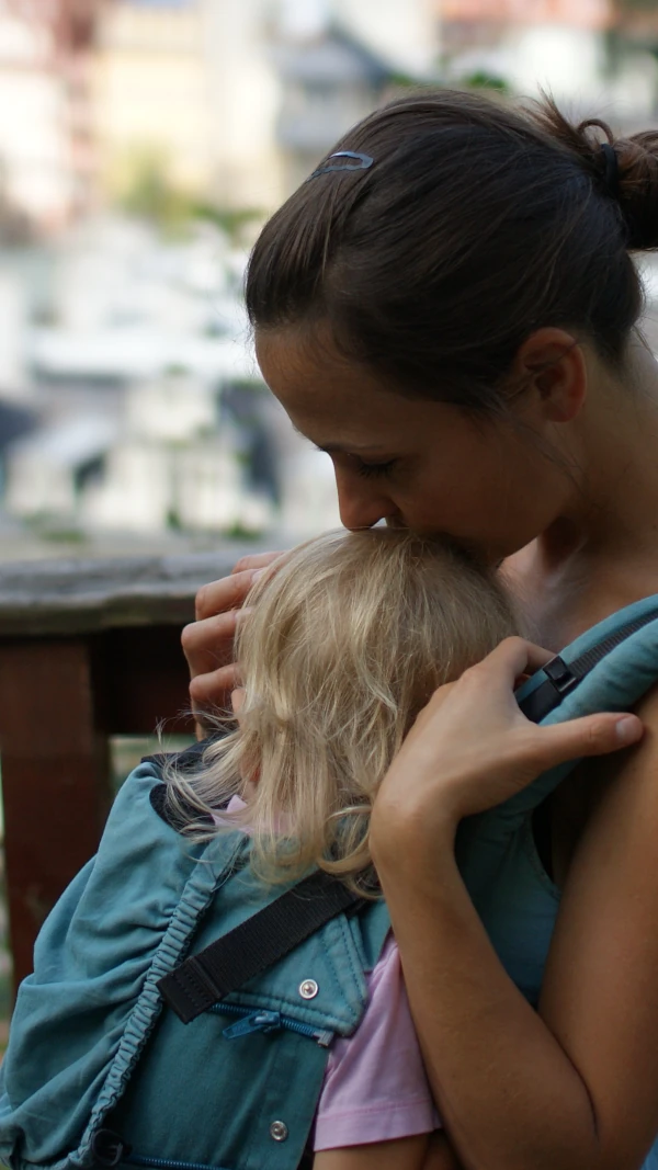 Eine Mutter (Evelin) hält ihr Kind in einer Babytrage und küsst es sanft auf den Kopf. Beide sind eng aneinander geschmiegt, das Kind ist nicht erkennbar.