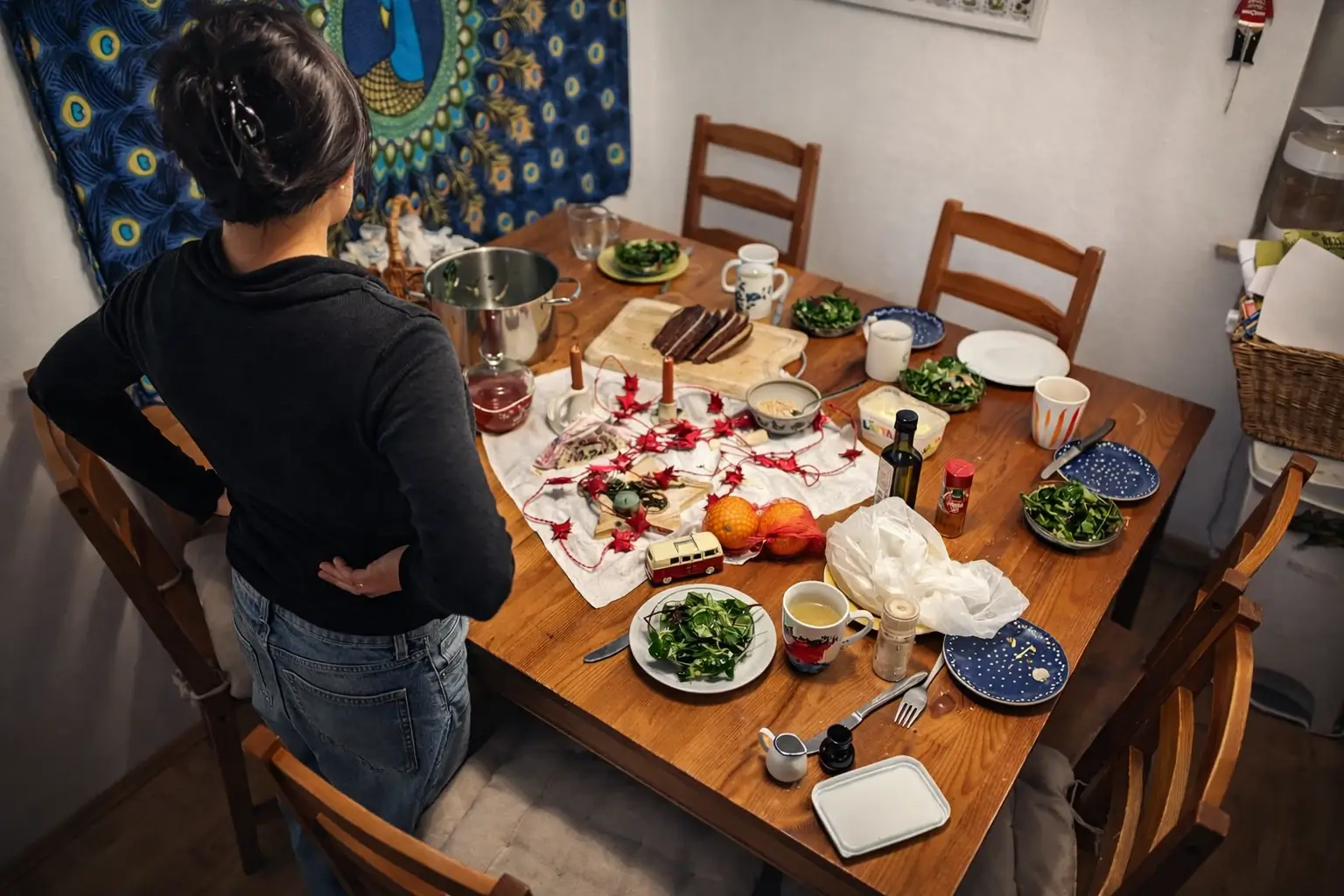 Eine Person steht an einem gedeckten Holztisch und blickt auf ein vorbereitetes Abendessen mit Salaten, Brot, Kerzen, Obst und Alltagsgegenständen in einer wohnlichen Küche.