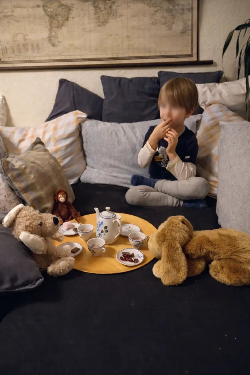 Ein Kind sitzt auf einem Sofa vor einem kleinen Tablett mit Spielzeug-Teeservice und teilt eine ruhige Kaffeerunde mit mehreren Kuscheltieren.
