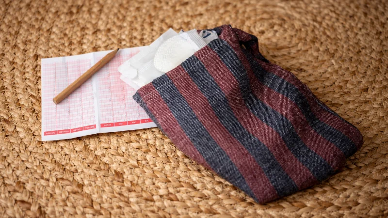 Kleines Menstruationsset mit Kalender, Binden und Stofftäschchen auf geflochtenem Teppich.