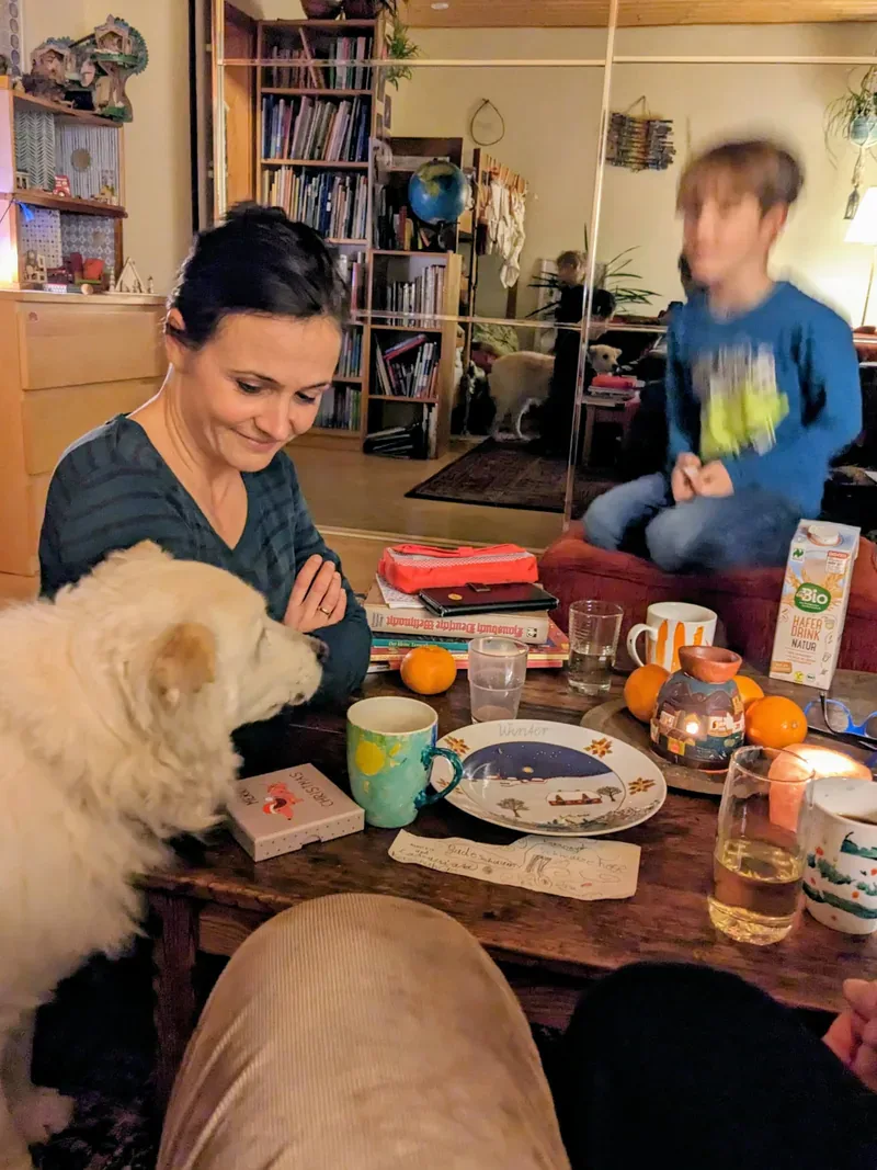 Eine erwachsene Person und ein Kind an einem gedeckten Tisch; ein Hund steht daneben.